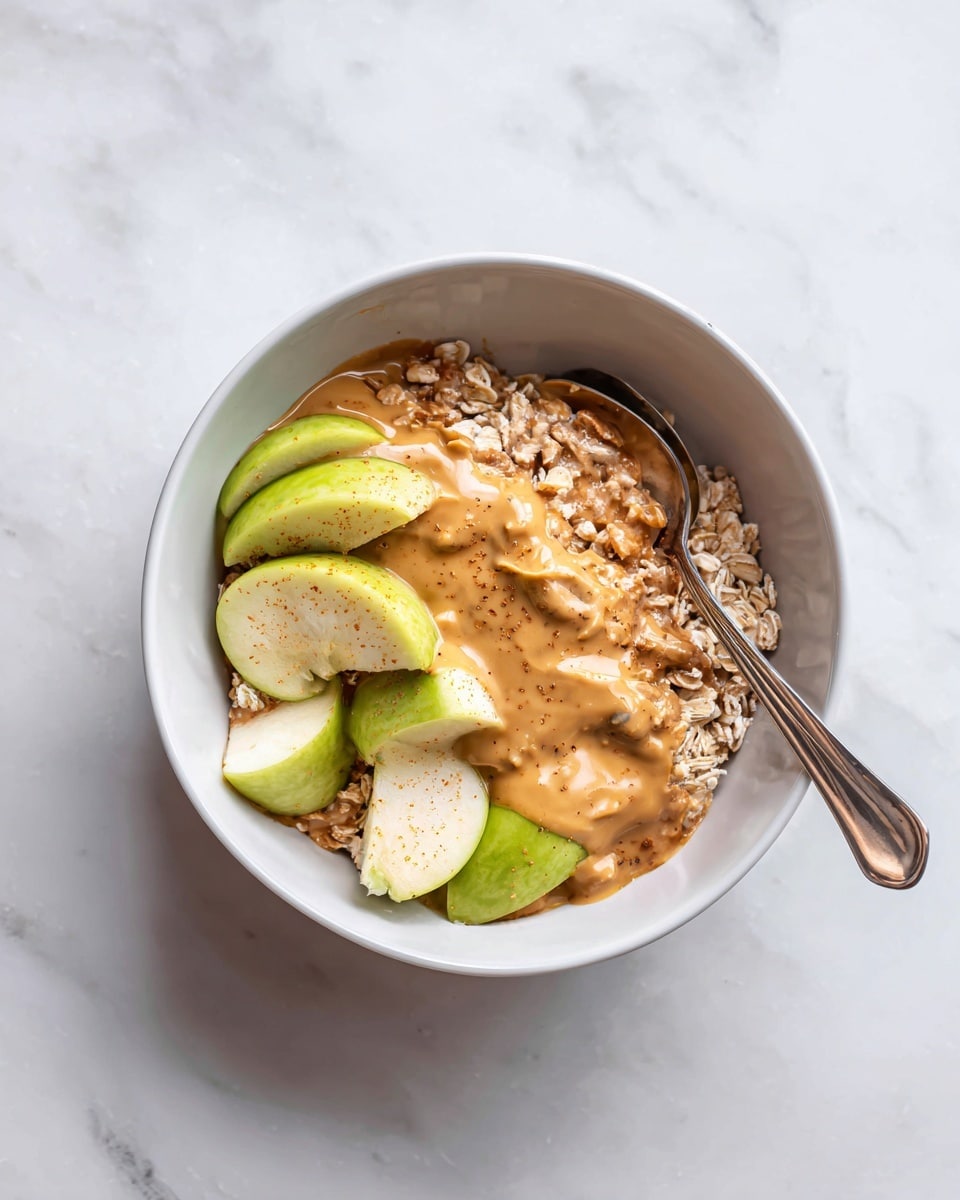 A white bowl on a white marbled surface contains a layered dish with three main layers. The bottom layer is light brown rolled oats with a rough texture, mostly covered but visible around the edges. The middle layer has several pieces of green apple with a shiny, smooth skin and white inside, placed unevenly on top of the oats. The top layer is light brown peanut butter sauce with a creamy and slightly thick texture, spreading over the apple pieces and oats, with some small bubbles visible. A spoon rests partly inside the bowl on the left side. Photo taken with an iphone --ar 4:5 --v 7