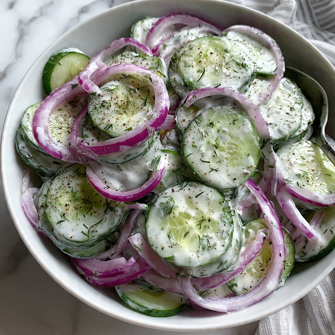 square image A white bowl filled with a fresh cucumber salad showing thin, round cucumber slices layered and mixed with thin rings of purple onion. The cucumber and onion layers are coated lightly with a creamy white dressing and sprinkled with small green dill herb pieces and ground black pepper. A metal spoon is partially visible in the bowl, resting to the side. The bowl sits on a white marbled surface, with a soft-focus striped cloth adding contrast at the edge. photo taken with an iphone --ar 4:5 --v 7