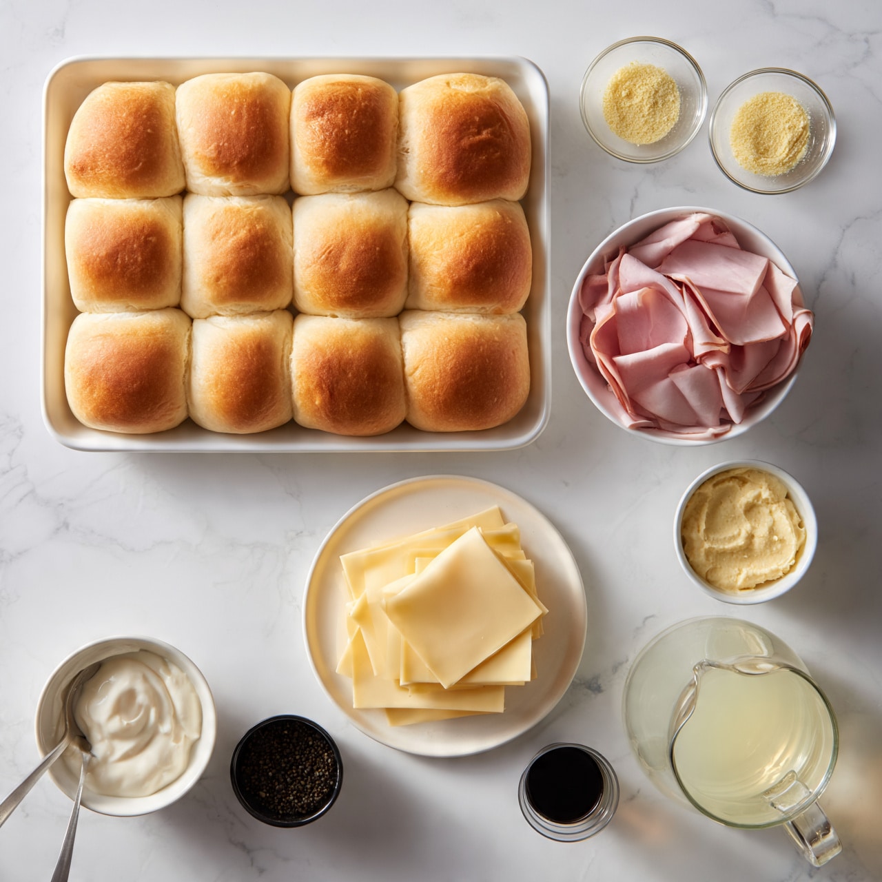 square image A white baking tray filled with 12 soft, golden brown dinner rolls arranged in a 3 by 4 grid at the top left side of the image; two small clear bowls with light yellow spices or powders are next to the top right; below, a white bowl holds thin slices of pink deli meat; a small white bowl of pale yellow spread is placed near it; in the center, a white plate contains several pale yellow cheese slices stacked neatly; at the bottom left is a small white bowl of a creamy white sauce with a spoon, near it a small black bowl with dark seeds; at the bottom right there is a clear glass pitcher filled with light yellow liquid and a small clear cup with dark liquid; all items are arranged on a white marbled surface photo taken with an iphone --ar 4:5 --v 7square image
