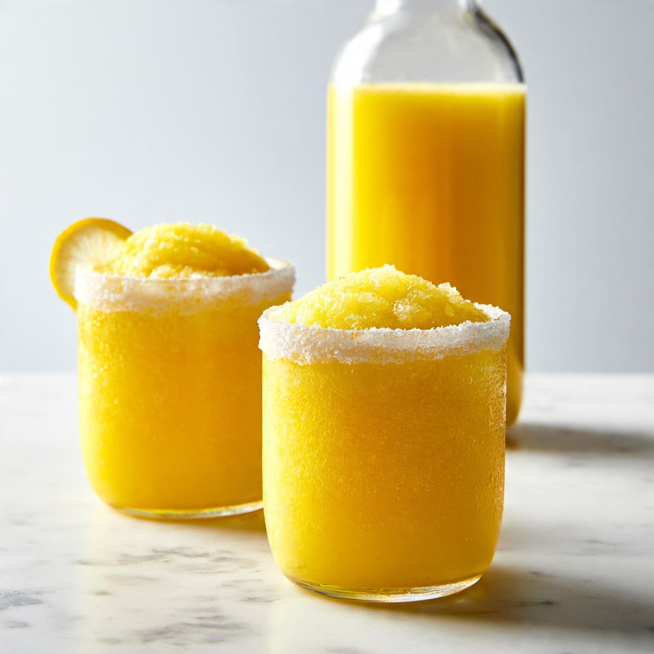 square image The image shows two short, round glasses filled with bright yellow slushy drinks. Each glass has a thick layer of icy texture on top, and the rims are coated with granulated sugar or salt. One glass has a small slice of lemon placed on its rim. Behind the glasses, there is a tall, clear bottle filled with the same yellow liquid. The setting features a white marbled surface and a simple, bright background. The photo taken with an iphone --ar 4:5 --v 7