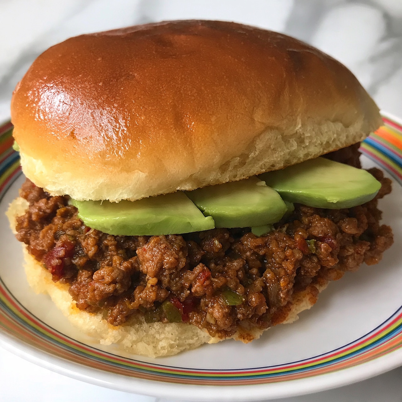 square image A close-up of a sandwich with a shiny, slightly toasted brown bun on top. Inside, there is a thick layer of cooked ground meat mixed with small pieces of red and green, likely vegetables or sauce, giving a chunky and moist texture. On top of the meat layer, there are three slices of light green avocado arranged side by side. The sandwich is placed on a white round plate with a colorful striped pattern visible around the edges. The background is a white marbled surface. Photo taken with an iphone --ar 4:5 --v 7