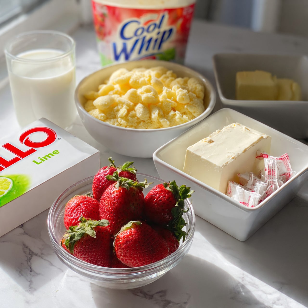 square image The image shows several ingredients on a white marbled surface. There is a white bowl filled with smooth, crushed yellow cookies in the center. To the right is a white square bowl with a block of cream cheese. Next to it, there are small wrapped sticks of butter. On the left side, there is a glass cup of white milk and a white box of Jell-O lime gelatin. A clear glass bowl full of fresh red strawberries with green leafy tops is in the front. Behind the bowls, a container of Cool Whip sits with a colorful label facing forward. A woman's hand is not seen interacting but the arrangement suggests preparing a dessert. The lighting is bright and natural. photo taken with an iphone --ar 4:5 --v 7