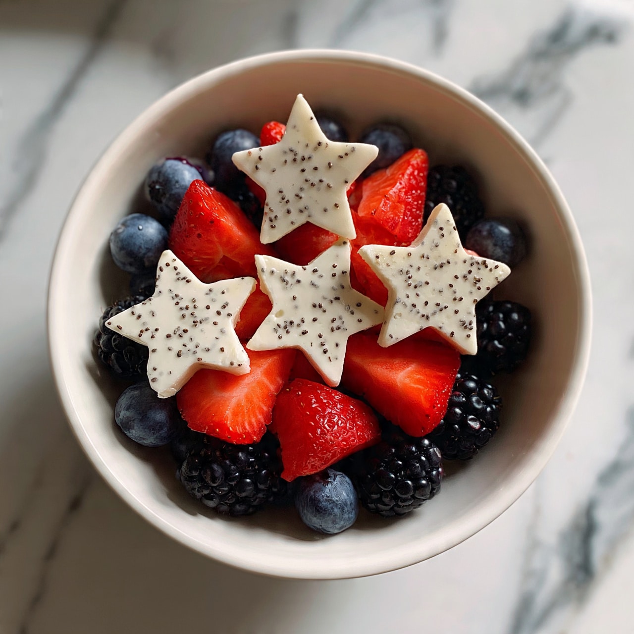 square image A white bowl filled with many small blueberries and blackberries as the base layer, topped with bright red strawberry pieces cut into triangles, and three large white star-shaped pieces sprinkled with tiny black seeds placed evenly on top, all set on a white marbled surface. photo taken with an iphone --ar 4:5 --v 7