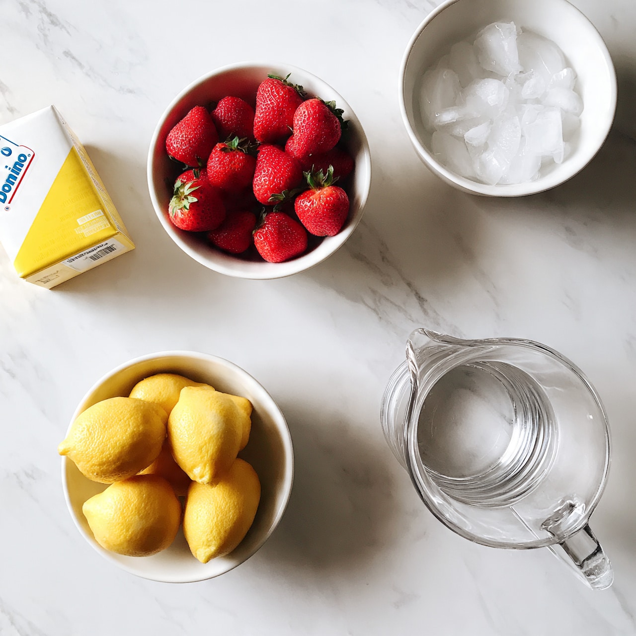 square image The image shows five items arranged on a white marbled surface: a box of Domino sugar with a yellow and white design on the left; a white bowl filled with bright red strawberries with green tops near the top center; a white bowl with ice cubes near the top right; a white bowl containing five yellow lemons near the bottom left; and a clear glass pitcher filled with water near the bottom right. The setup is simple and clean, with bright colors from the fruit standing out against the light background. photo taken with an iphone --ar 4:5 --v 7