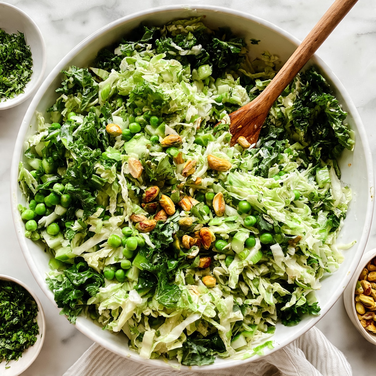 square image A large white bowl is filled with a fresh salad containing multiple layers of shredded light green cabbage, darker green kale leaves, small bright green peas, and light green cucumber pieces. Scattered on top are golden brown pistachio nuts and small green herb leaves. A wooden spoon stands inside the bowl, mixing the salad. Around the bowl, there is a small white bowl with green chopped herbs and another small bowl filled with more pistachio nuts. The whole scene is set on a white marbled surface with a light striped cloth near the bowl. photo taken with an iphone --ar 4:5 --v 7