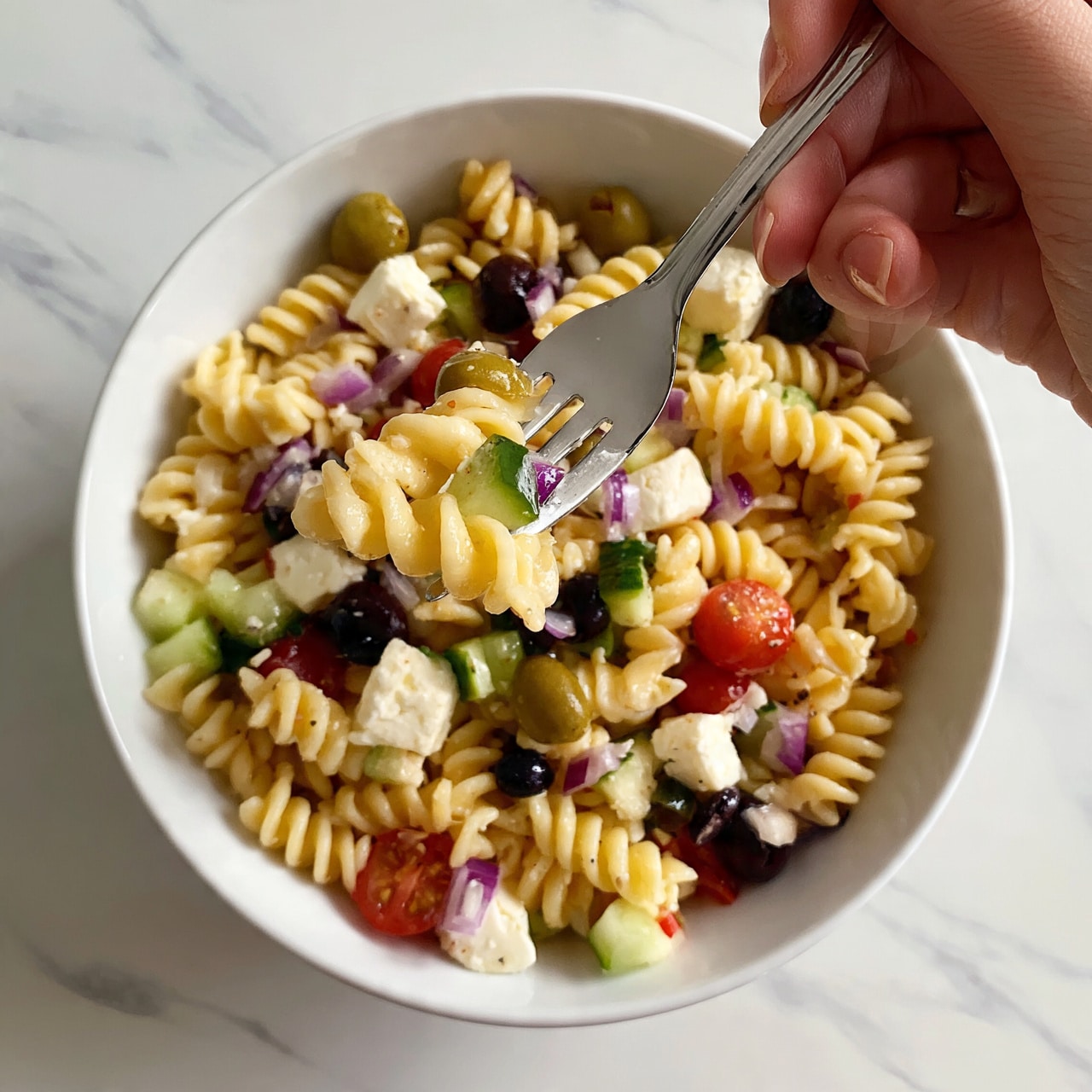 square image The dish shows a white bowl full of spiral pasta mixed with small pieces of cucumber, cherry tomatoes, green and black olives, purple onion, and cubes of white cheese. The pasta is light yellow and the vegetables add bright colors like green, red, and purple. A woman's hand holds a silver fork that lifts some pasta and vegetables out of the bowl. The bowl is on a white marbled surface. The photo taken with an iphone --ar 4:5 --v 7