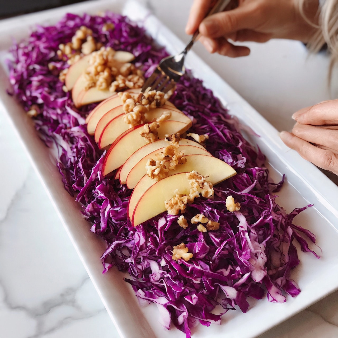 square image A long white plate holds a fresh salad with three layers. The bottom layer is shredded purple cabbage, filling most of the plate with a bright texture. On top, thin slices of red apple with pale yellow skin are spread evenly across the cabbage. Small walnut pieces are scattered over the salad, adding extra texture and a light brown color. The salad is placed on a white marbled surface, with a woman’s hand holding a fork near the plate. The photo taken with an iphone --ar 4:5 --v 7