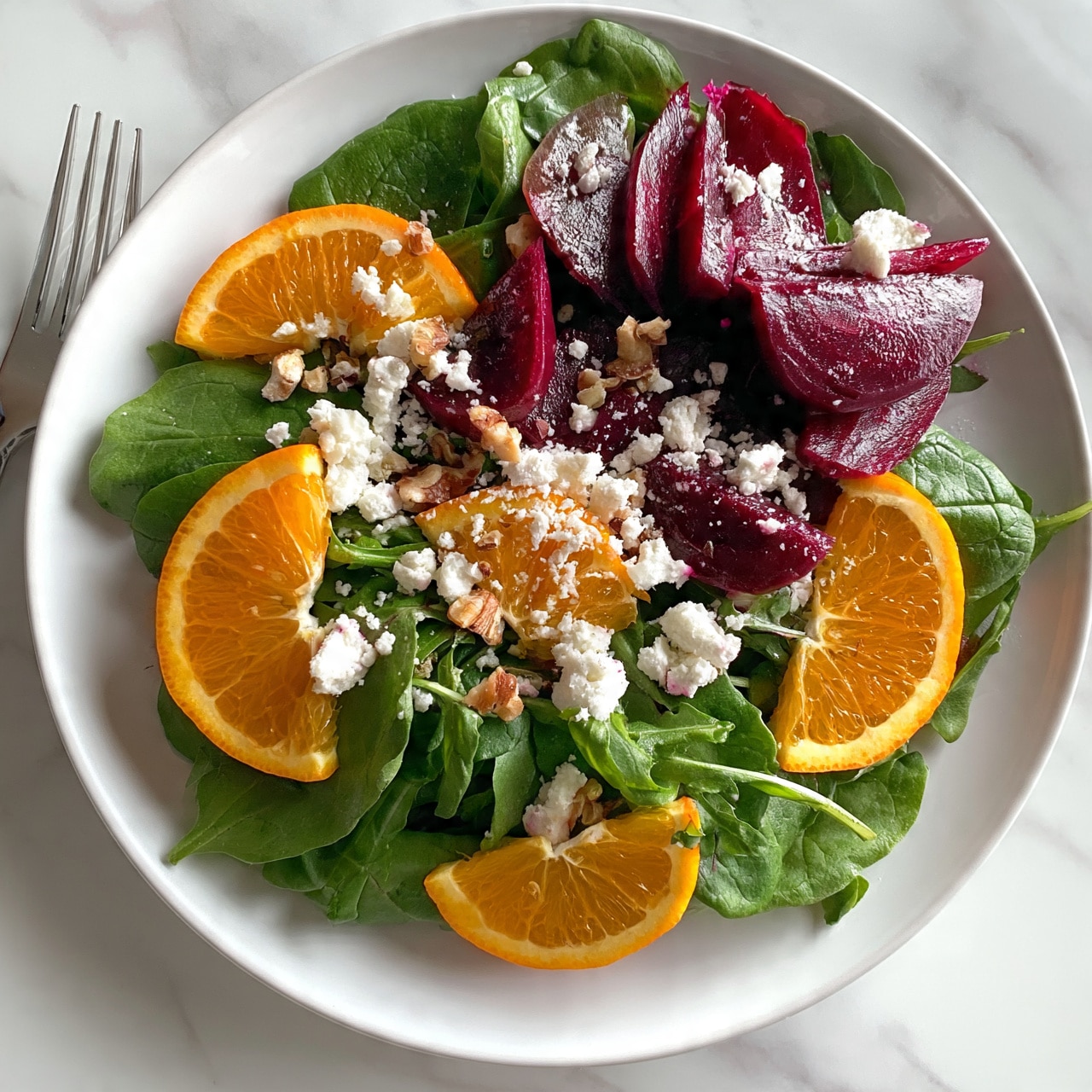 square image A white plate holds a colorful salad with three main layers visible. The bottom layer is fresh green spinach and arugula leaves spread evenly across the plate. On top of the greens, there are bright orange slices placed around the edges and a few in the middle, adding a pop of vibrant color. The top layer consists of deep red and purple beet slices, some folded and layered slightly on one side of the plate. Crumbled white cheese is sprinkled over the salad, along with small walnut pieces scattered gently across the top. The plate is on a white marbled surface, and a silver fork is placed to the left side. The photo appears natural and well-lit, capturing the freshness of the ingredients. photo taken with an iphone --ar 4:5 --v 7