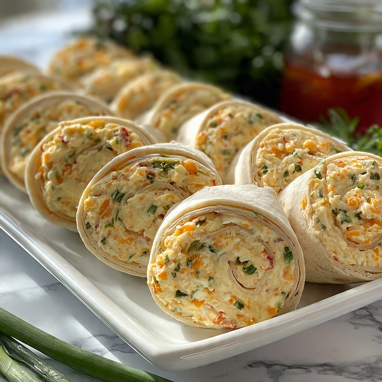 square image The image shows a white rectangular plate filled with several round pinwheel sandwiches arranged in neat rows. Each sandwich has a creamy, pale yellow filling mixed with small pieces of orange, green, and red, suggesting ingredients like cheese, herbs, and vegetables. The outer layer is a soft, light-colored tortilla or flatbread, rolled tightly around the filling. In the background, there are fresh green herbs and a clear glass jar with a reddish liquid. The plate is placed on a white marbled surface with some green onions lying nearby. photo taken with an iphone --ar 4:5 --v 7
