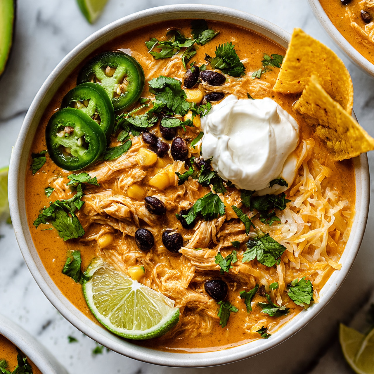square image A white bowl filled with a creamy orange soup layered with shredded chicken and scattered black beans and corn, topped with thin slices of bright green jalapeño on the left side, fresh green cilantro leaves sprinkled throughout, a dollop of thick white sour cream near the center, two yellow tortilla chips leaned on the right edge, shredded cheddar cheese mixed in, and a fresh lime wedge resting on the bottom left of the bowl, all placed on a white marbled surface with parts of avocado, lime wedges, and another bowl of soup visible around it photo taken with an iphone --ar 4:5 --v 7
