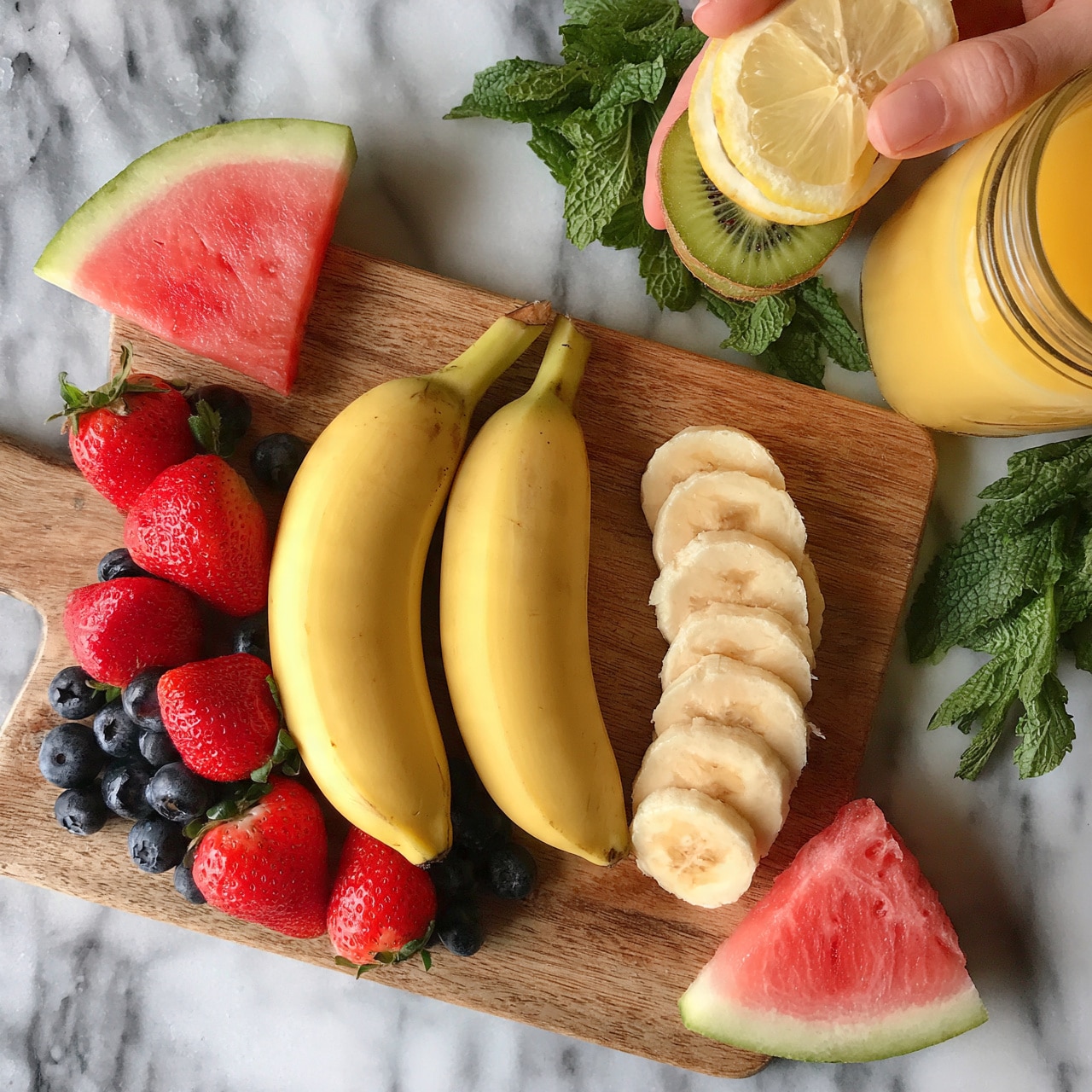 square image The image shows a wooden board on a white marbled surface, topped with two whole yellow bananas and one banana peeled in slices arranged neatly on the right side of the board. There are bright red strawberries and dark blueberries scattered around the board for color contrast, with two slices of pink watermelon placed at the top left and bottom right corners of the board. To the right of the board, a woman’s hand is holding a stack of citrus slices including lemon and kiwi, next to a glass jar filled with orange juice and fresh green mint leaves scattered nearby. Photo taken with an iphone --ar 4:5 --v 7