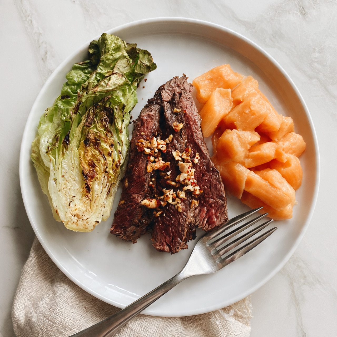 square image A white plate on a white marbled surface holds a meal with three main parts: on the left, a small wedge of grilled green lettuce with visible char marks, in the center, three thick slices of dark reddish-brown meat topped with some crushed nuts or seeds, and on the right, a portion of soft, light orange carrots. A silver fork rests on a beige cloth napkin partially under the plate. The photo taken with an iphone --ar 4:5 --v 7