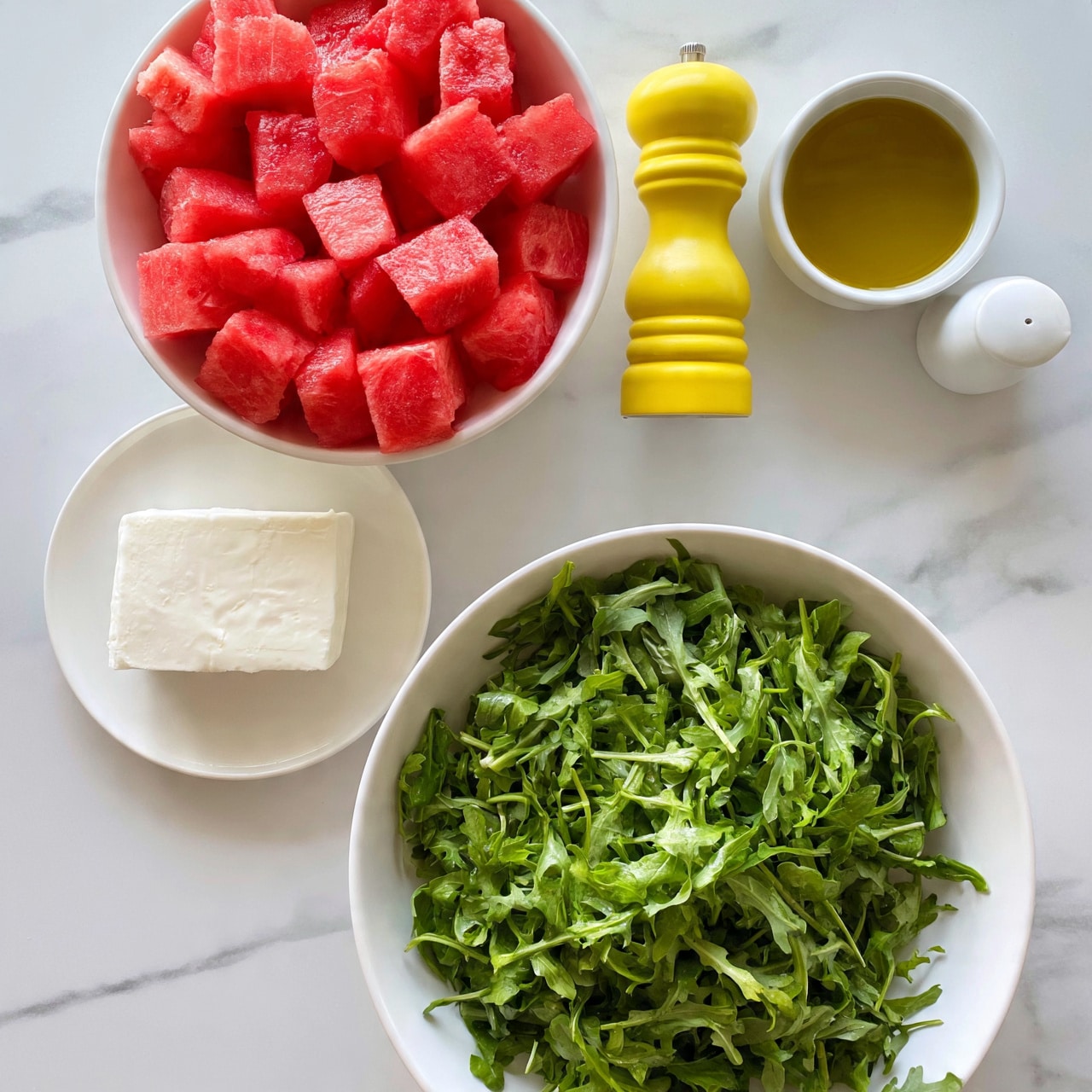 square image The image shows a top view of fresh salad ingredients arranged separately on a white marbled surface. In a large white bowl at the bottom right, there is a big pile of green arugula leaves with a fresh, leafy texture. To the left, on a white plate, there is a block of white cheese with a smooth texture. Above it, a white bowl is filled with bright red watermelon cubes, each with a juicy and slightly grainy texture. To the right of the watermelon, there are three items in a row: a yellow pepper grinder with a smooth surface, a white small bowl filled with golden olive oil, and a white salt shaker. The whole setup is clean and colorful. Photo taken with an iphone --ar 4:5 --v 7