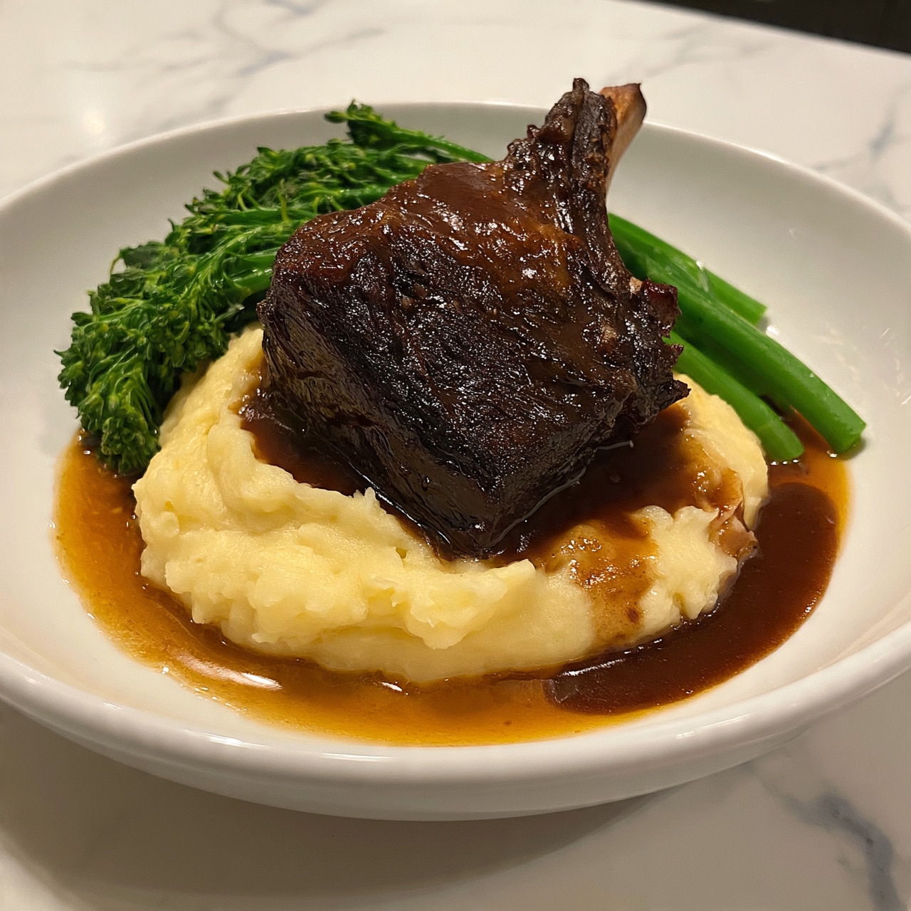 square image The image shows a white plate with three layers of food. At the bottom is a smooth, creamy layer of mashed potatoes with light yellow color. On top of the mashed potatoes rests a large, dark brown cooked meat piece with a bone sticking out, covered with a thick brown sauce that also pools around the mashed potatoes. Next to the meat and potatoes, there are bright green broccolini stalks arranged neatly. The plate is on a white marbled surface. photo taken with an iphone --ar 4:5 --v 7