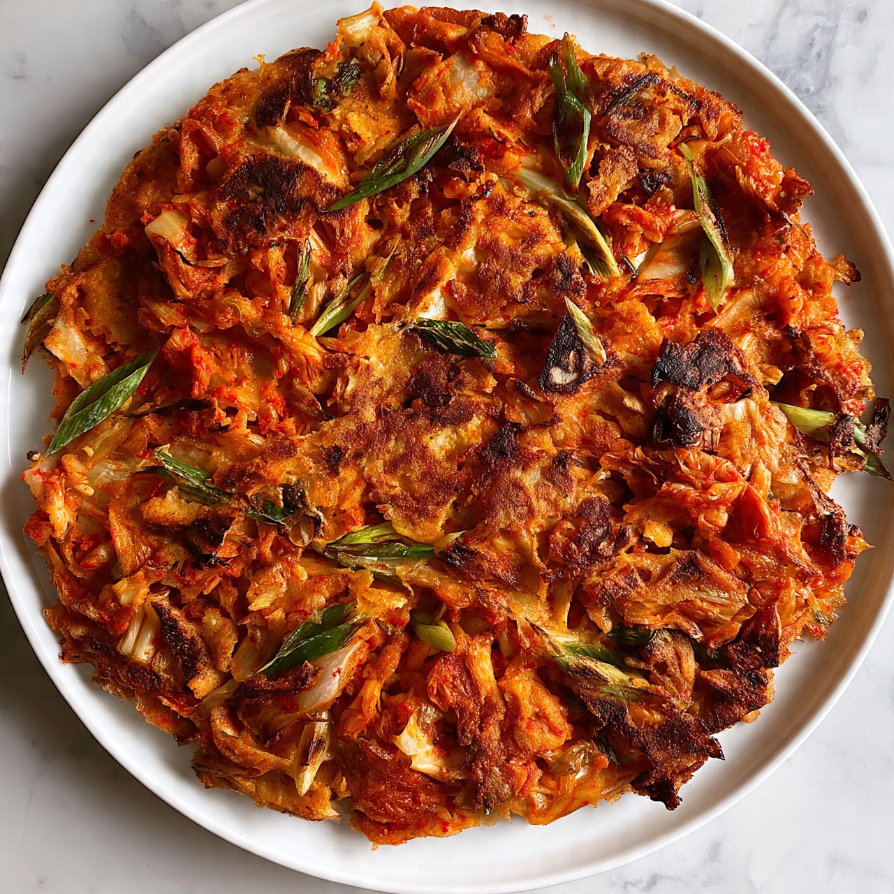 square image A round, flat kimchi pancake with a crispy, golden-brown surface mixed with bright red-orange kimchi pieces and green scallion slices scattered throughout, all placed on a white plate on a white marbled surface photo taken with an iphone --ar 4:5 --v 7
