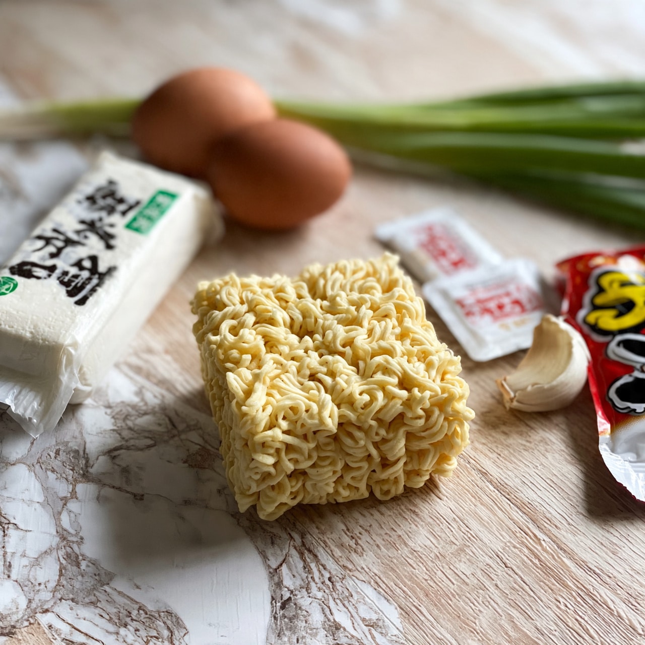 square image The image shows several ingredients for a meal placed on a light wooden surface with a white marbled texture added. At the center is a block of yellow, dry instant ramen noodles with a wavy texture and square shape. To the left, there is a white tube of silken tofu laying sideways, with clear writing in black and green on the label. Behind the tofu is a brown egg and a long green stalk of spring onion. To the right side of the noodles, there are two small white seasoning packets with red and black text, placed next to a peeled clove of garlic and part of an open red packet bag with some yellow, black, and white lettering visible. The scene is simple with soft natural light, and there is no presence of any woman’s hand. Photo taken with an iphone --ar 4:5 --v 7