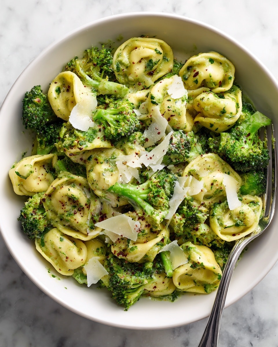 A white bowl filled with green tortellini pasta mixed with bright green broccoli florets, all coated in a light green sauce with visible herbs. On top, there are thin white cheese shavings scattered evenly across the dish. A shiny silver fork rests inside the bowl on the right side. The background is a white marbled surface. photo taken with an iphone --ar 4:5 --v 7