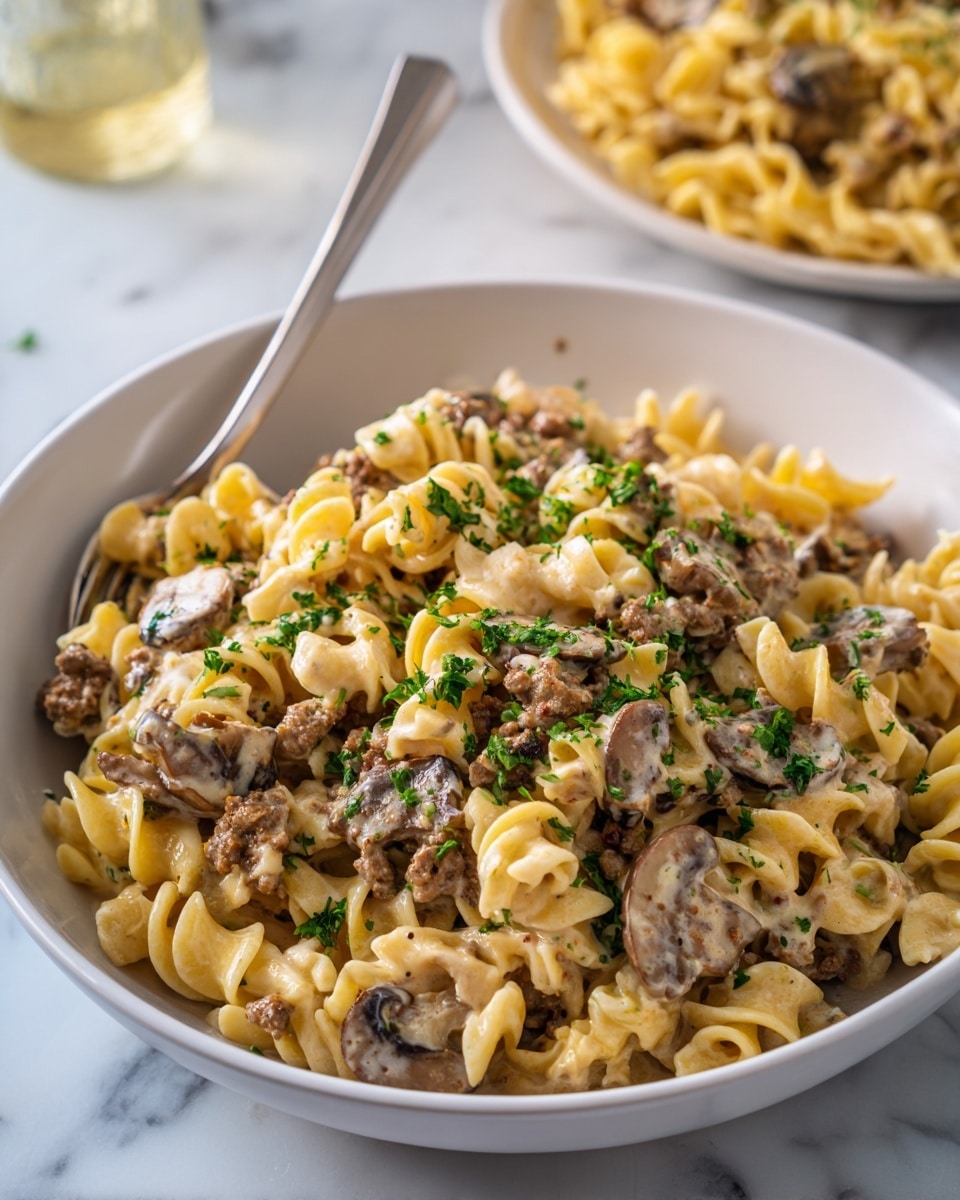 This image shows a large white bowl filled with a creamy pasta dish made of twisted egg noodles mixed with browned ground meat and sliced mushrooms. The creamy sauce covers the noodles and meat evenly, giving a smooth texture. The dish is topped with small sprinkles of fresh green chopped parsley, adding a touch of bright color. A metal spoon rests inside the bowl on the left side, ready for serving. In the background, there is a blurred white plate filled with the same pasta, all placed on a white marbled surface. photo taken with an iphone --ar 4:5 --v 7