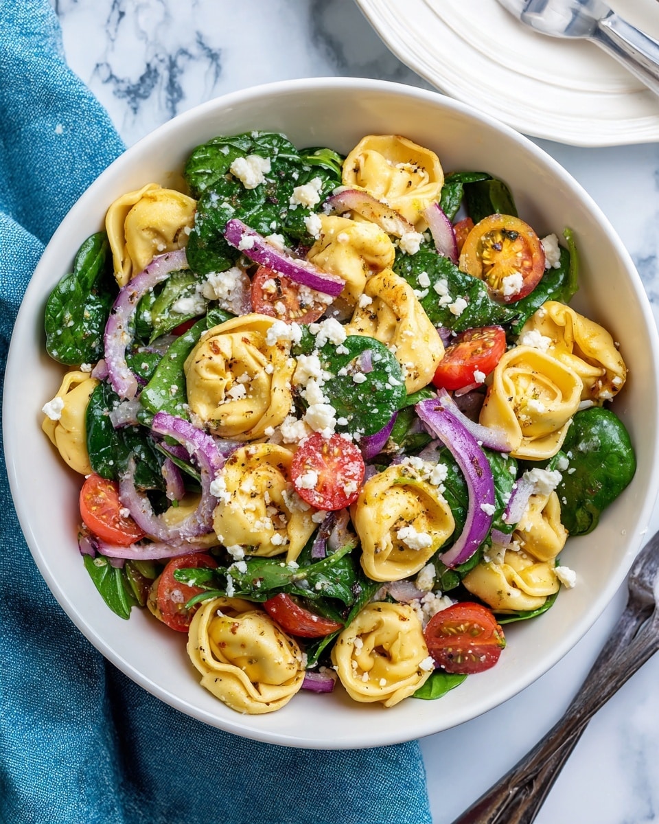 A white bowl filled with tortellini pasta mixed with fresh green spinach leaves, bright red cherry tomato halves, and small pieces of purple onion. The tortellini are golden with a slightly browned texture, and the salad is sprinkled with white crumbled cheese. The bowl sits on a white marbled surface with a blue cloth underneath, alongside a white plate with a fork and knife resting on it. Photo taken with an iphone --ar 4:5 --v 7
