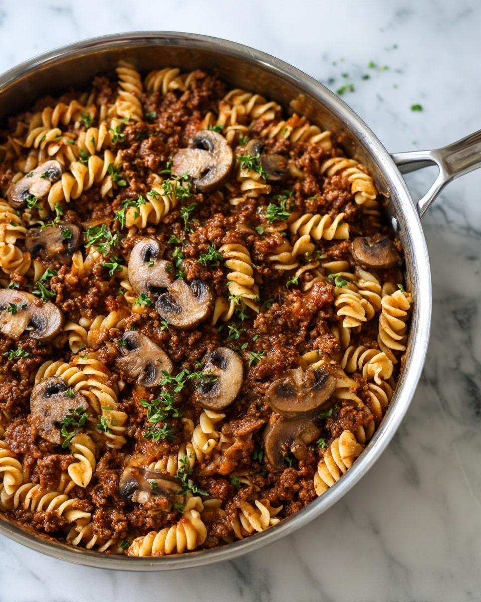 This image shows a close-up of a cooked beef and mushroom pasta dish in a silver pan. The dish is made of three main layers: the bottom layer consists of cooked ground beef with a brown, slightly saucy texture; the middle layer includes mushroom slices with a soft, tender texture and light brown color mixed evenly throughout; the top layer features short curly noodles coated in a rich brown sauce, dispersed evenly over the beef and mushrooms. Small green herb pieces are sprinkled across the surface. The pan is placed on a white marbled surface. photo taken with an iphone --ar 4:5 --v 7