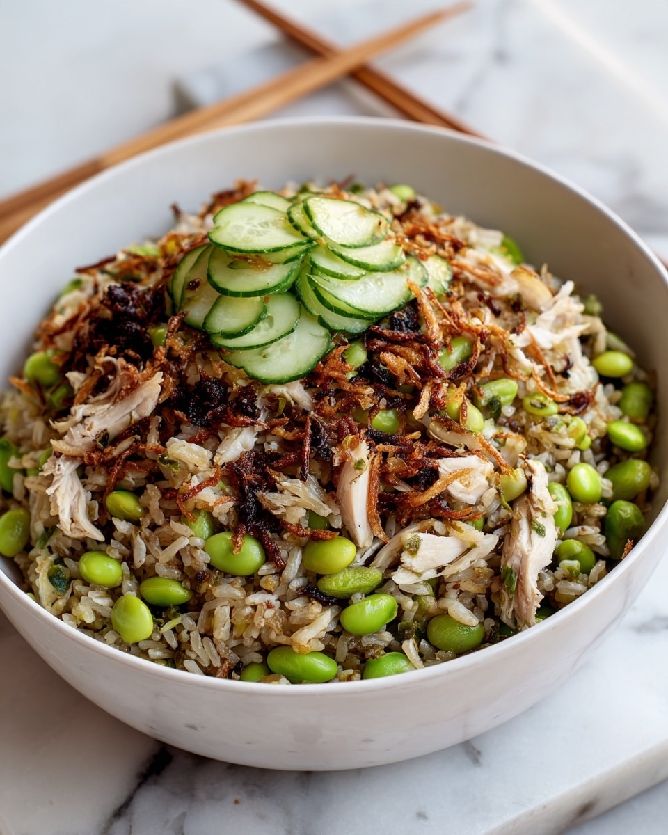 A white bowl filled with a layered rice dish where the bottom layer is light brown rice mixed with green edamame beans scattered throughout and thin slices of cucumber on top. The next layer has shredded light brown and white pieces of cooked chicken mixed with small dark seeds and bits of crispy golden-brown fried onions. The dish has a slightly glossy texture with some charred darker bits adding contrast. The bowl sits on a white marbled surface with a pair of light wooden chopsticks nearby. Photo taken with an iphone --ar 4:5 --v 7