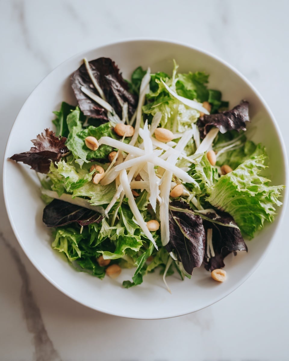 The image shows a fresh salad in a white bowl, filled with different layers. The base layer is made of bright green leafy lettuce and some darker purple leaves mixed in. On top of this, there are long, thin white strips that look soft and light. Scattered around the salad are small, pale brown nuts or seeds that add texture. The salad is placed on a white marbled surface with soft natural light. photo taken with an iphone --ar 4:5 --v 7