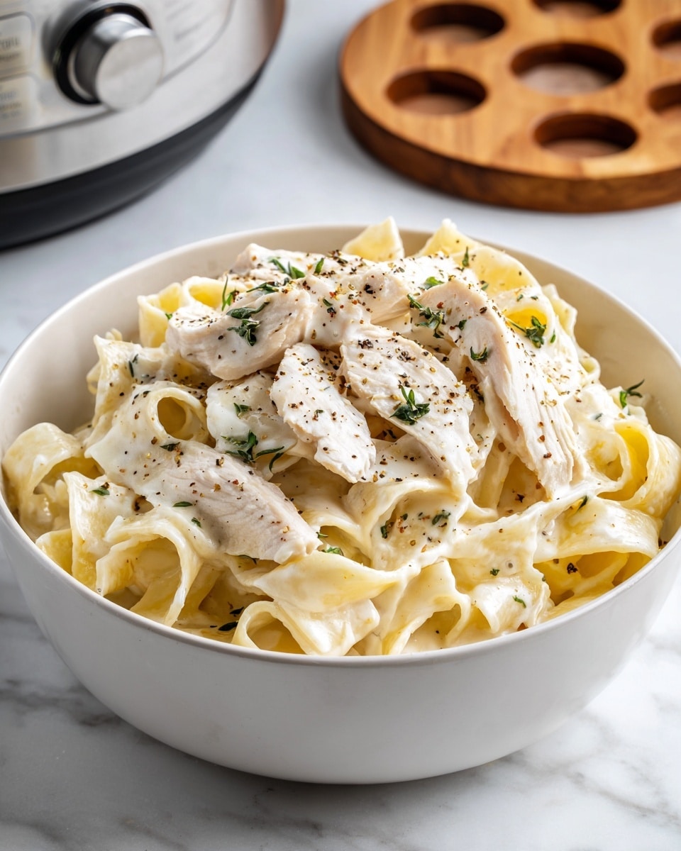 A white bowl filled with creamy pasta that has wide, flat noodles covered in a thick white sauce. On top are several strips of white chicken mixed into the pasta, with small green chopped herbs sprinkled evenly over the dish. A light dusting of black pepper is also visible across the surface. The bowl is placed on a white marbled surface with a round wooden trivet with three slots in the background near a metallic appliance knob. photo taken with an iphone --ar 4:5 --v 7