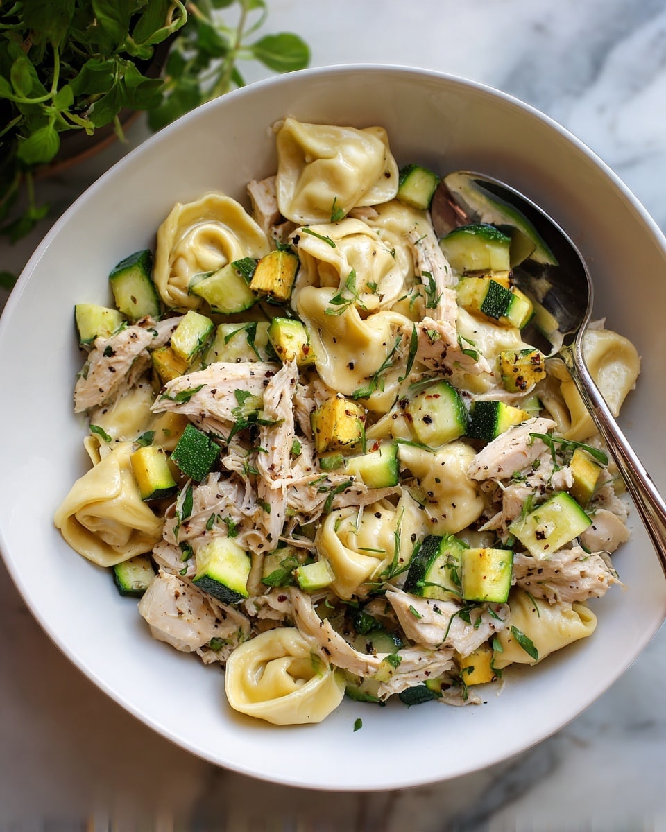 A white bowl filled with a mix of three layers: pale yellow tortellini pasta with a soft, slightly wrinkled texture, light beige shredded chicken pieces, and bright green diced zucchini cubes with dark green skin. The ingredients are spread evenly, with pasta and chicken pieces tangled together and zucchini scattered throughout, all sprinkled with small black seasoning bits. A silver spoon rests inside the bowl, reflecting light softly. The bowl sits on a white marbled surface with some green plants in the blurred background photo taken with an iphone --ar 4:5 --v 7