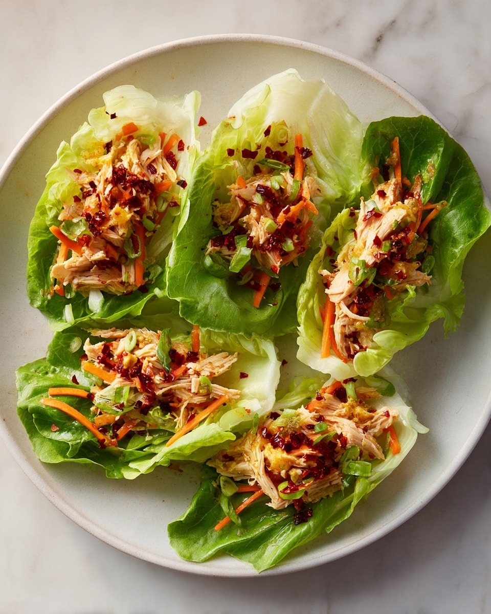 Five green lettuce leaves lie flat on a large round white plate. Each leaf is filled with a mix of light beige shredded chicken, thin orange carrot strips, and small light yellow pieces, all sprinkled with dark red chili flakes. The texture of the lettuce is crisp and fresh, with some leaves folded slightly at the edges. The plate rests on a white marbled surface with soft natural lighting highlighting the fresh colors. photo taken with an iphone --ar 4:5 --v 7