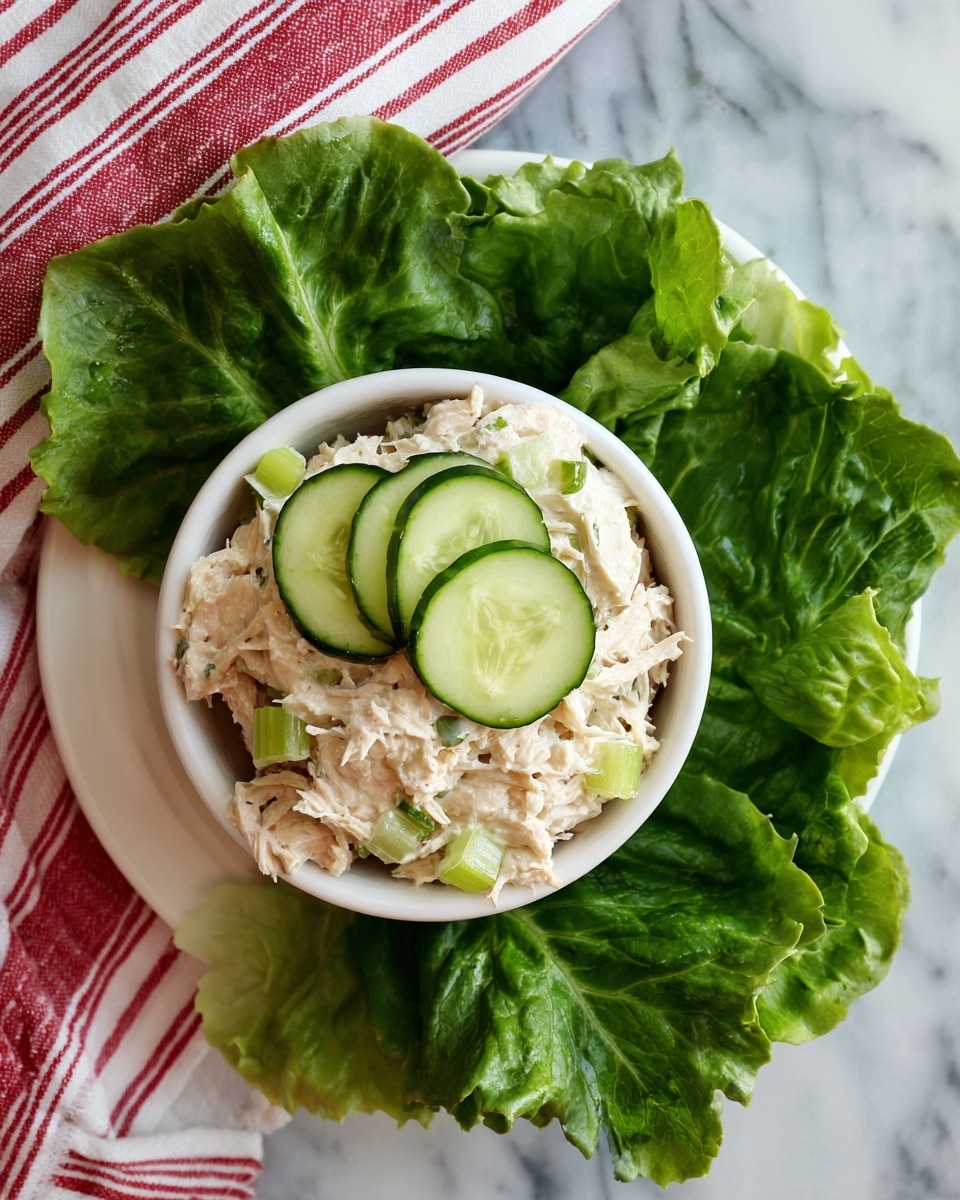 A white bowl filled with a creamy mixture of shredded light-colored tuna and chopped light green celery pieces, topped with three round cucumber slices with dark green edges in the center; the bowl rests on a white plate and is surrounded by fresh green lettuce leaves beneath, placed on a white marbled surface with a red and white striped cloth partially visible underneath, photo taken with an iphone --ar 4:5 --v 7