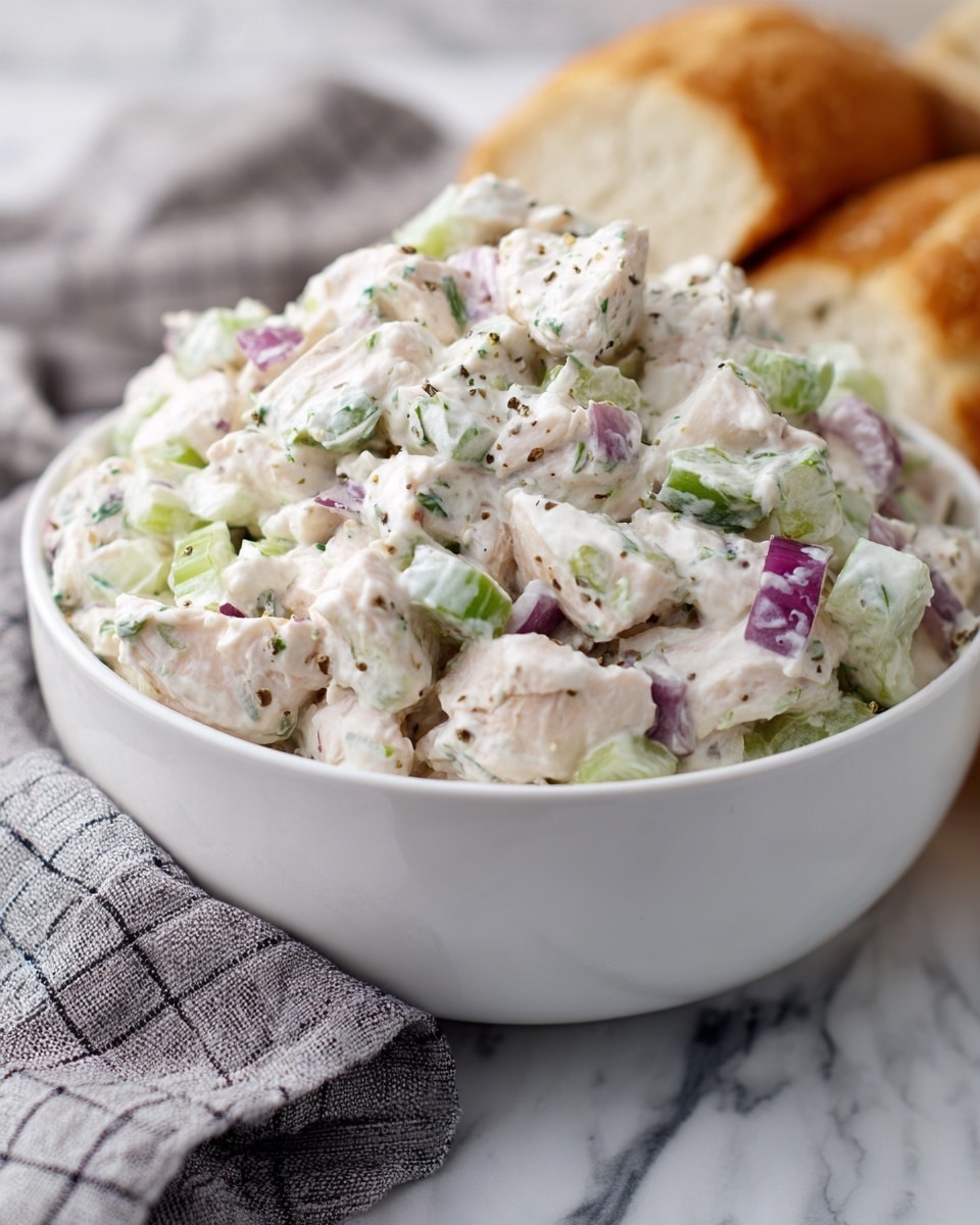 A white bowl filled with a creamy chicken salad that has visible chunks of white chicken, light green celery pieces, and small slices of purple-red onion mixed together with a thick, smooth white dressing. The bowl sits on a white marbled surface with a soft gray checkered cloth partially visible on the left side and blurred bread pieces in the background on the right. The salad looks fresh and chunky with a mix of smooth and crunchy textures. Photo taken with an iphone --ar 4:5 --v 7