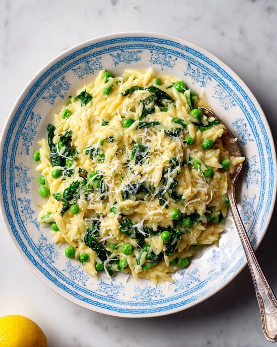 A white plate with a light blue pattern around the edge holds a dish with three main layers: the bottom layer is creamy light yellow orzo pasta, mixed softly with bright green peas and bits of dark green spinach, spread evenly. On top, there is a sprinkling of finely grated pale yellow cheese that looks soft and crumbly. A silver spoon rests slightly submerged in the pasta, ready to scoop a bite. On the white marbled surface next to the plate, there is a small part of a yellow lemon in the corner. The image shows the food clearly with natural light, making the colors fresh and bright, photo taken with an iphone --ar 4:5 --v 7