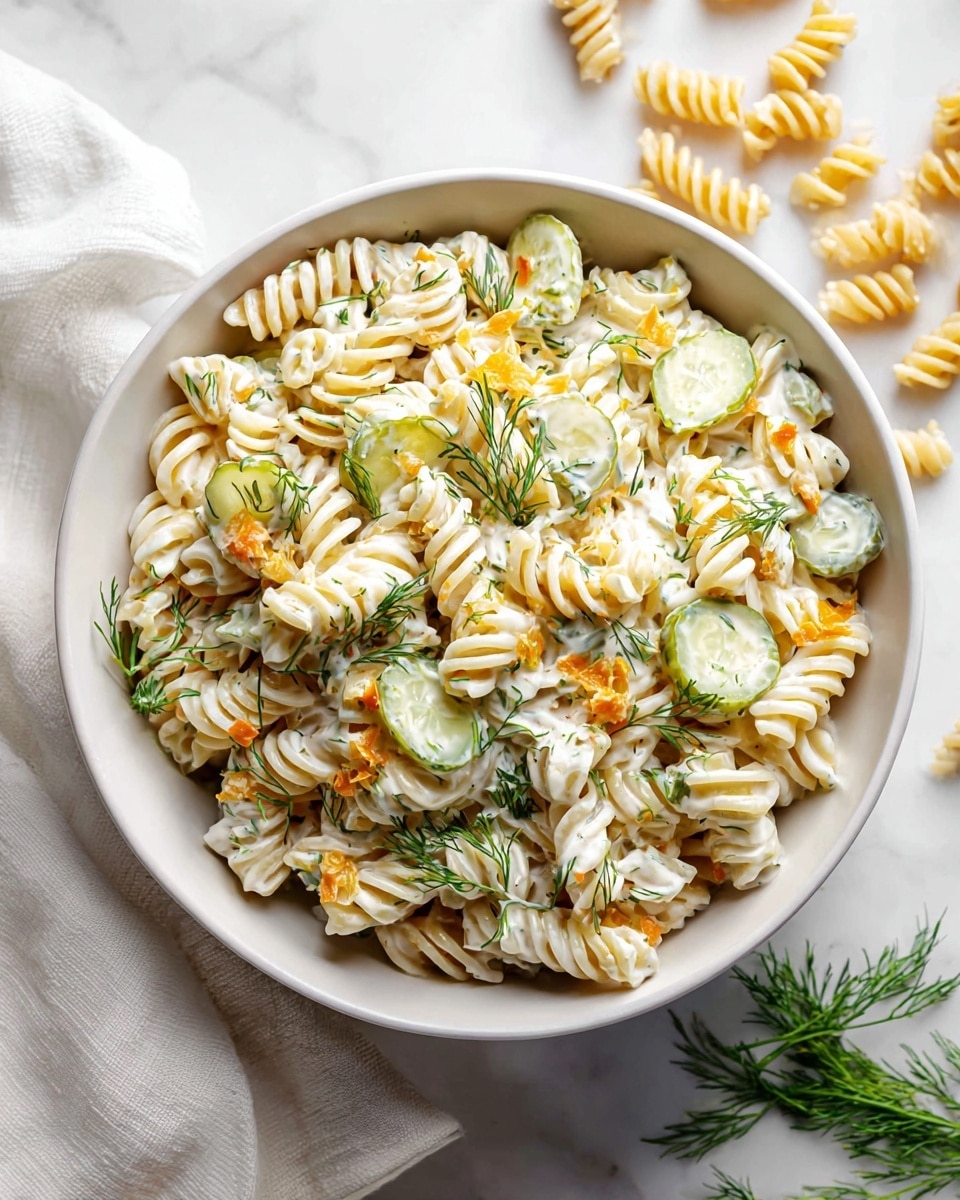 A white bowl filled with rotini pasta mixed with creamy white dressing and small orange cheese cubes. There are thick slices of green pickles scattered on top and around the pasta. Fresh dill is sprinkled lightly over the dish, adding a touch of green color. The bowl is set on a white marbled surface with a few dry rotini pasta pieces beside it, and a white cloth is placed near the bowl. Photo taken with an iphone --ar 4:5 --v 7