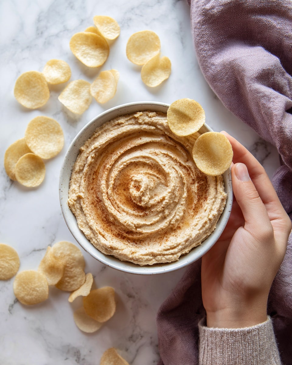 The image shows a white ceramic bowl filled with a light brown creamy dip with swirls of a slightly darker color mixed in. Two round, pale yellow chips are lightly placed on top in the center of the bowl. Around the bowl, there are more chips scattered on a white marbled surface. A woman's hand holds the edge of the bowl from the left side. The background has a soft purple cloth partially visible behind the bowl. Photo taken with an iphone --ar 4:5 --v 7