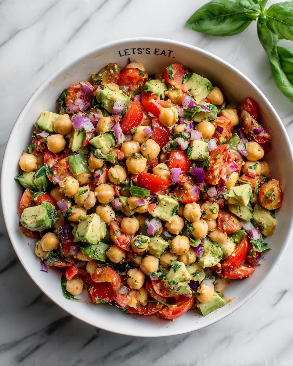 A bowl filled with a fresh chickpea salad, showing three main layers of ingredients mixed evenly: round beige chickpeas, bright red tomato chunks, and green avocado pieces with some darker green basil strips scattered throughout. Small bits of finely chopped purple onion are spread over the salad adding a pop of color and texture. The salad looks lightly coated with seasoning, giving a slight shine to the chickpeas and vegetables. All ingredients sit inside a white bowl with the words